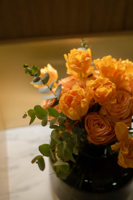 A close-up of a floral arrangement featuring vibrant orange ranunculus and roses, complemented by sprigs of silver-green eucalyptus, placed in a glossy black vase on a white surface. The arrangement d