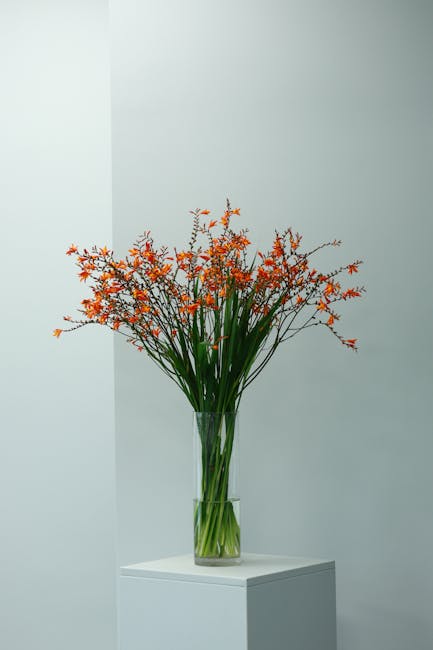 A tall, elegant glass vase filled with a large arrangement of fresh orange and green flowers, including long, slender green stems and vibrant orange blossoms, is placed on a white pedestal against a n