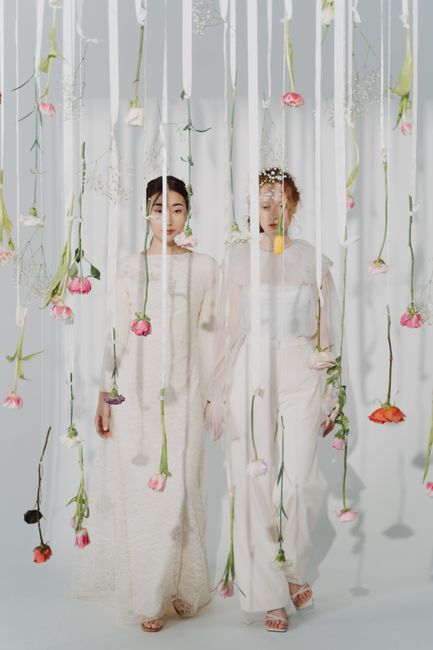 Two women stand behind a sheer white curtain decorated with hanging pink and peach roses with green stems, creating a floral backdrop. The women are dressed in elegant, light-colored dresses, with one