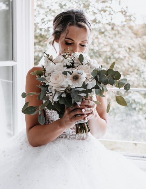 A young bride with fair skin and styled hair is holding a large, elegant bouquet of fresh flowers, including white roses, white anemones with dark centers, and various eucalyptus leaves. The bouquet i