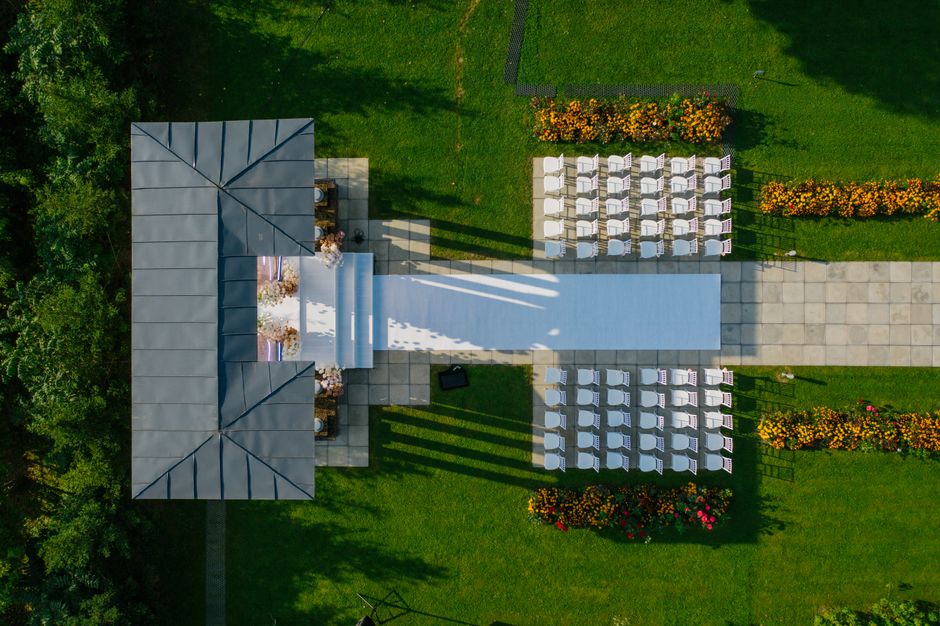 Aerial view of a wedding ceremony setup in a lush green outdoor area, featuring a white aisle runner leading to a decorated arch with floral arrangements, surrounded by neatly arranged white chairs fo